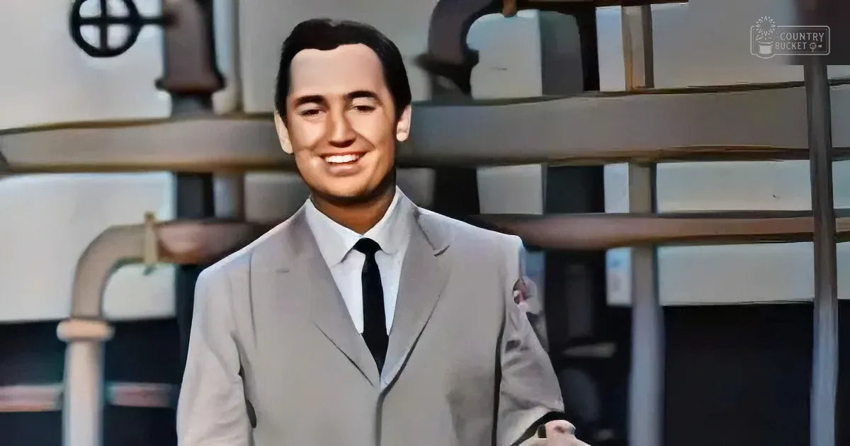 A man in a light gray suit and black tie smiles in front of a backdrop with pipes and machinery.