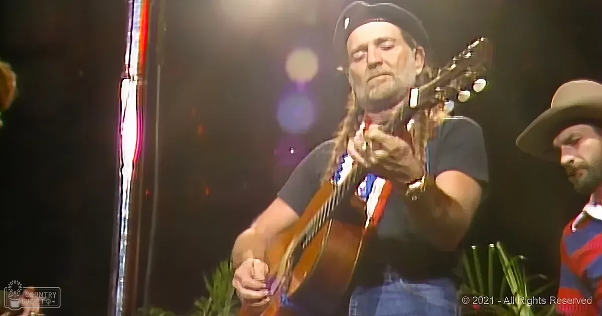 A man with long hair and a hat plays a guitar on stage, surrounded by other musicians.