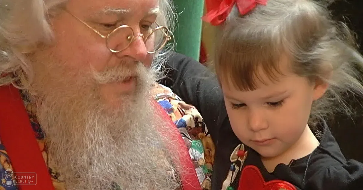 A toddler boy wearing a yellow and blue plaid shirt being hugged by an older adult dressed as Santa Claus in a home setting with a Christmas tree visible, indicating winter season.