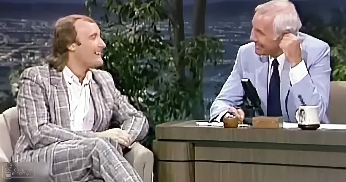 A man in a plaid suit sits on a couch, smiling, while another man in a suit leans forward, appearing to listen intently.