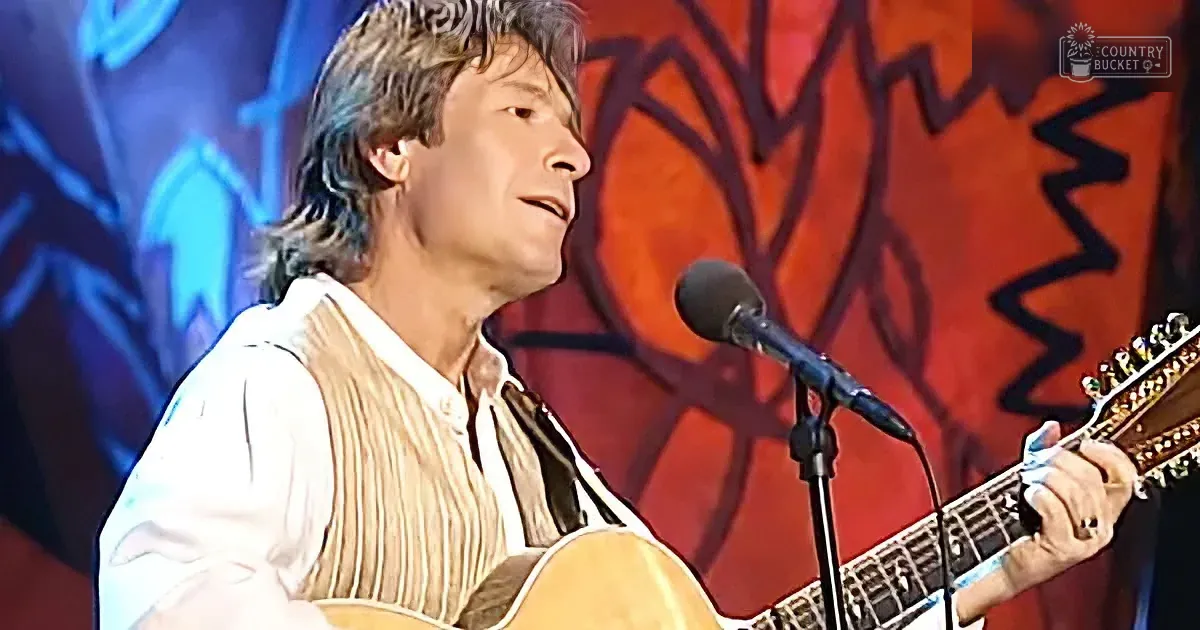 A man plays an acoustic guitar on stage, wearing a white shirt and vest, with a colorful background.