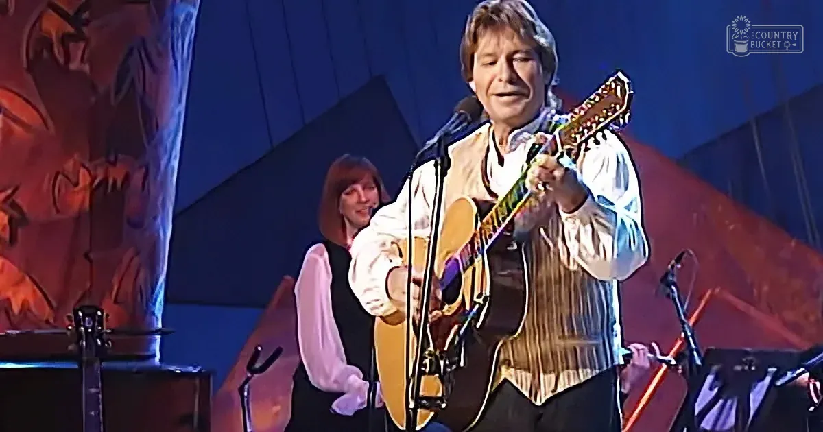 A man plays guitar on stage, singing into a microphone, with a woman in the background.