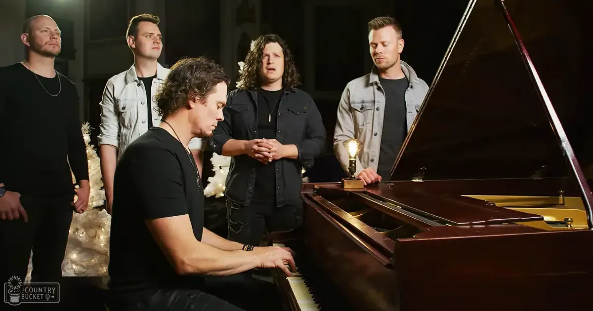 A man plays piano while four others watch, one with clasped hands, in a dimly lit room.