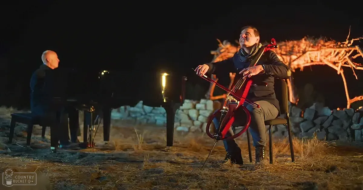 A man plays a red cello under a night sky, illuminated by a lantern, while another man plays a piano nearby.