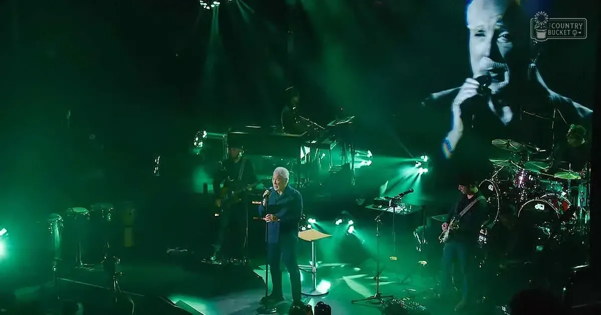 A man sings on stage with a band, illuminated by green lights, with a large screen showing a close-up of him.