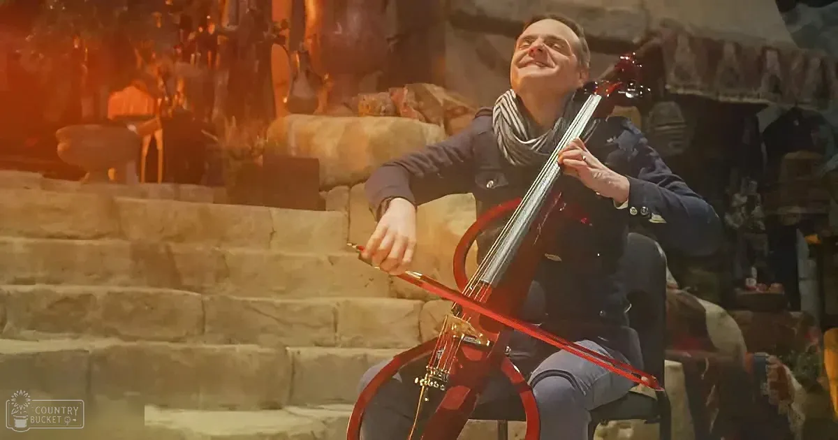 A man sits on stone steps, playing a red cello with a warm, glowing background.