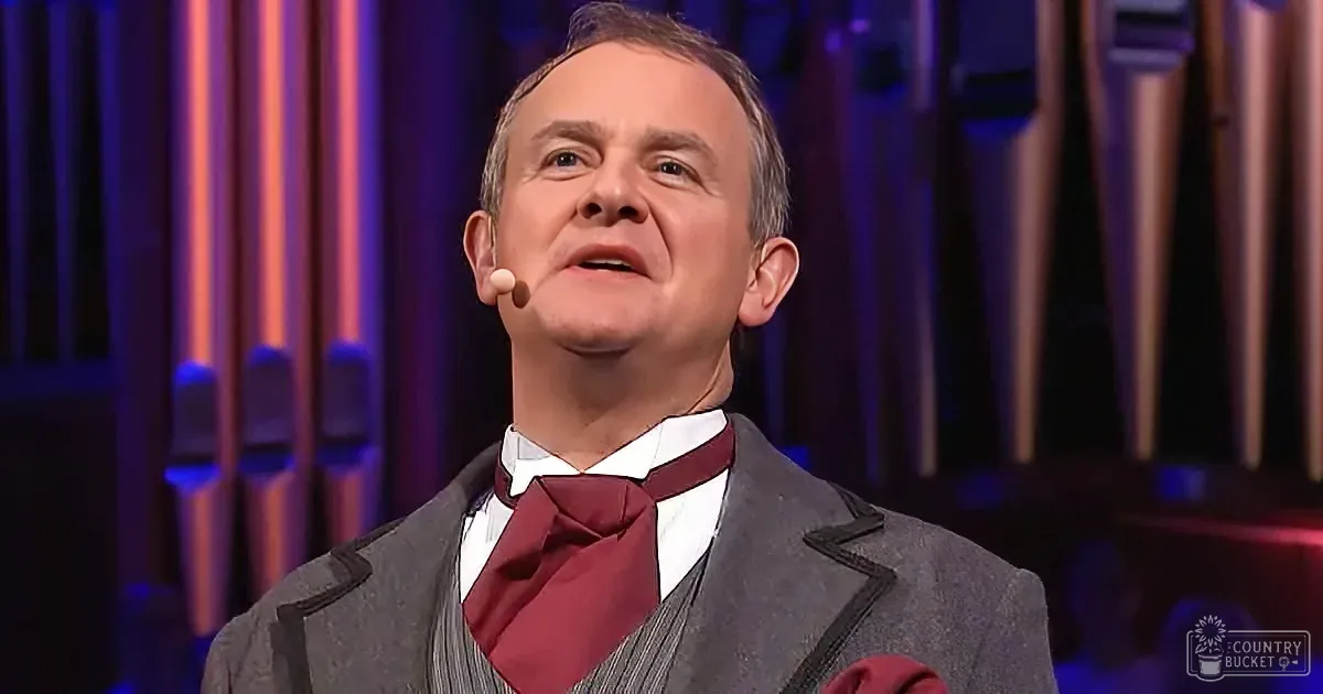 A man in a suit and bow tie stands on stage with a microphone, looking upwards.