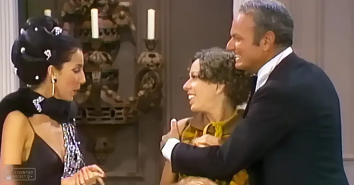 A man in a suit embraces a woman in a yellow dress, while another woman in a black dress looks on.