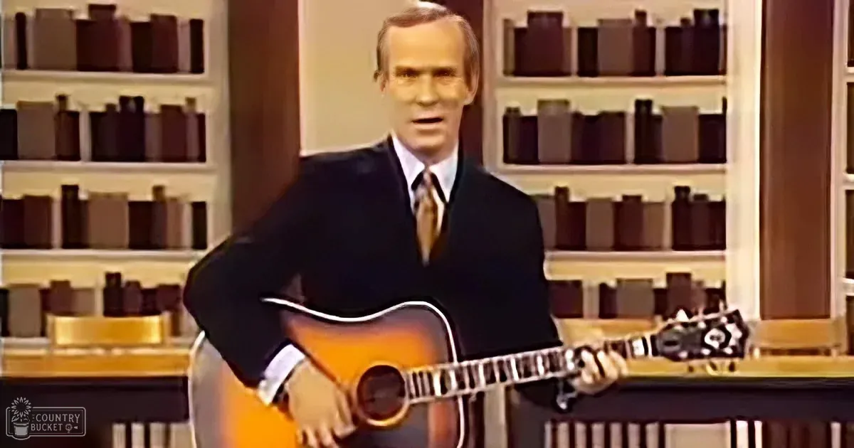 A man in a suit holds an acoustic guitar, standing in front of shelves filled with books.