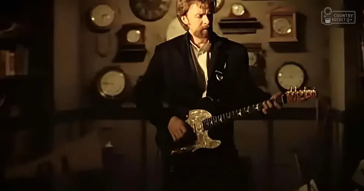 A man in a suit holds a guitar, standing in front of a wall with clocks.