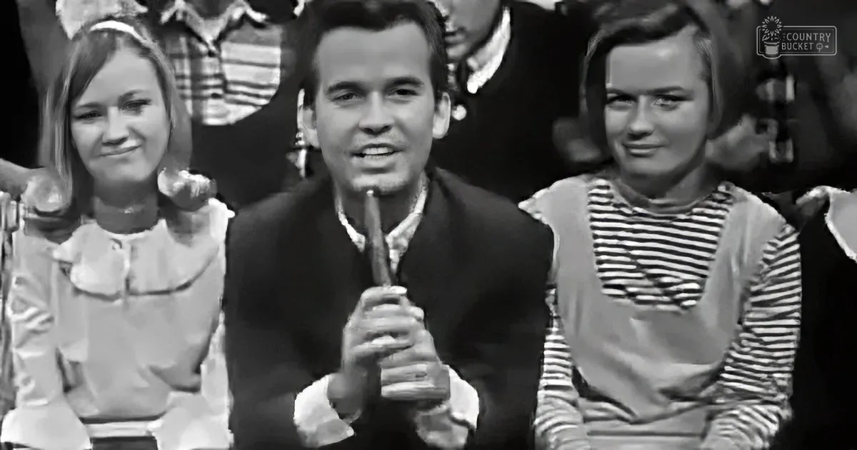 A man in a suit holds a microphone, flanked by two women in striped shirts, in a vintage setting.