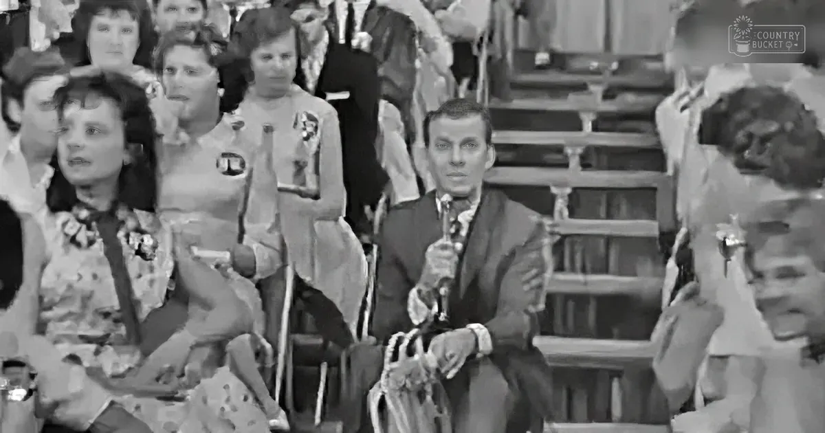 A man in a suit holds a microphone, seated in front of a crowd at a formal event.