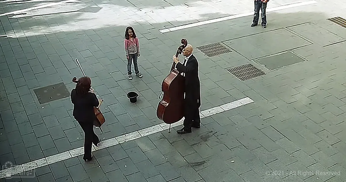 A man in a suit plays a cello in a public square, with a woman and another musician nearby.