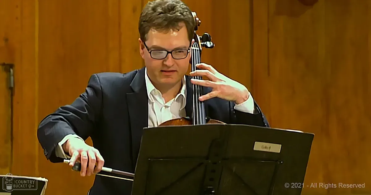 A man in a suit plays a cello, holding the bow and looking at sheet music.