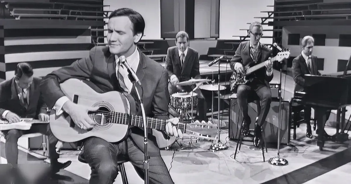 A man in a suit plays guitar on stage with a band, including a drummer, bassist, and pianist.
