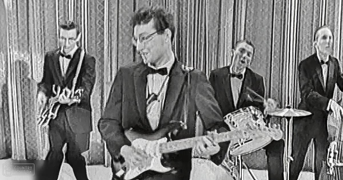 A man in a suit plays guitar while a saxophonist, drummer, and another musician in a tuxedo look on.
