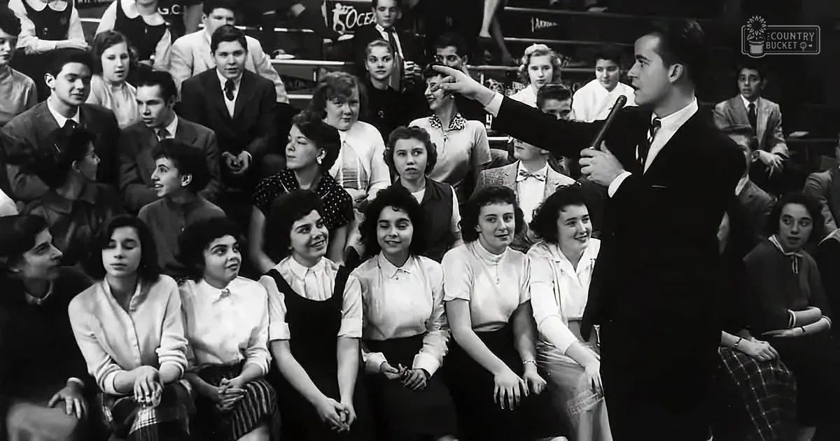 A man in a suit points at a seated audience of students in a gymnasium.