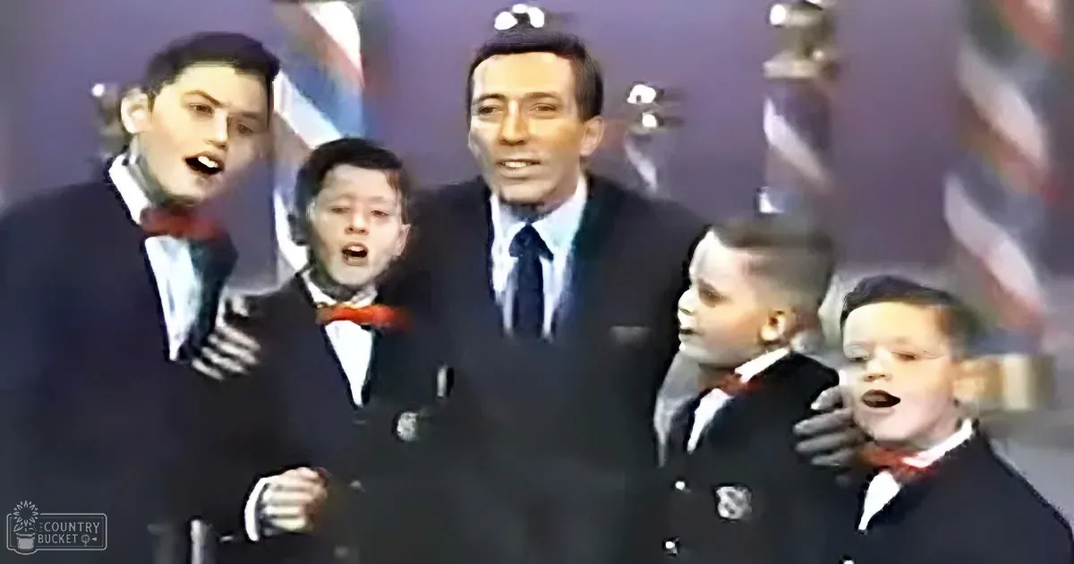 A man in a suit sings with four young boys in suits and bow ties, all appearing to perform.