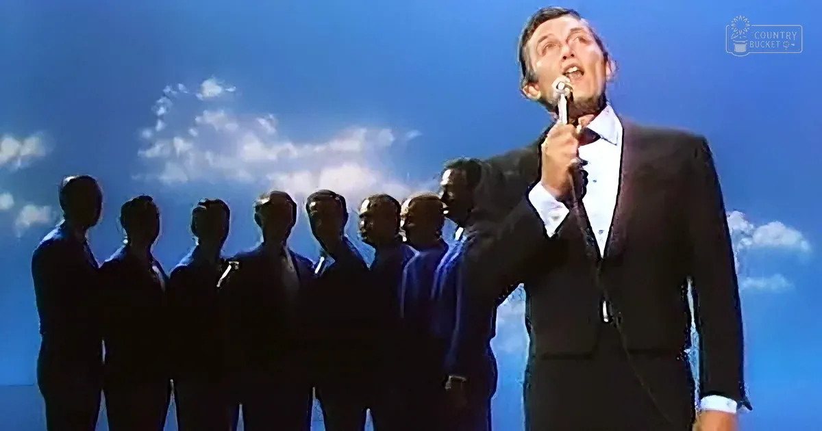 A man in a suit sings into a microphone, silhouetted against a blue sky with clouds.