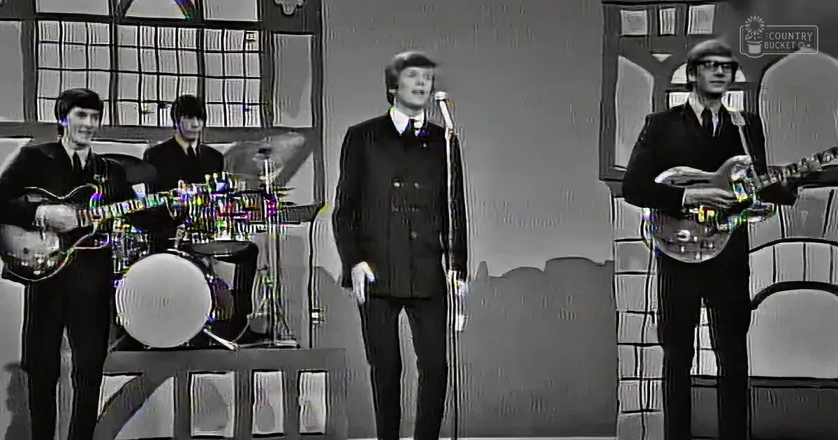 A man in a suit sings into a microphone, accompanied by a band playing guitars and drums.