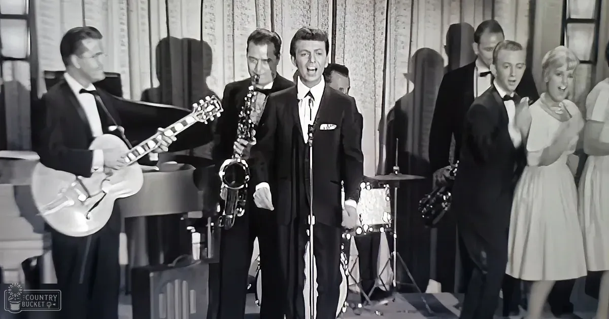 A man in a suit sings into a microphone, surrounded by musicians and a choir in formal attire.