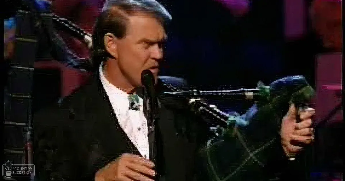 A middle-aged male performer wearing a dark suit jacket and white shirt with a tie plays green and black bagpipes in an indoor venue with dramatic purple lighting.