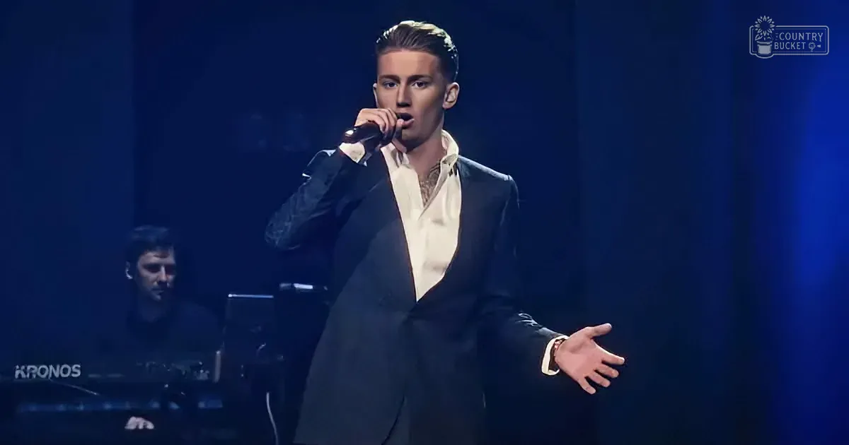 A man in a suit sings into a microphone on stage, with a keyboard player in the background.