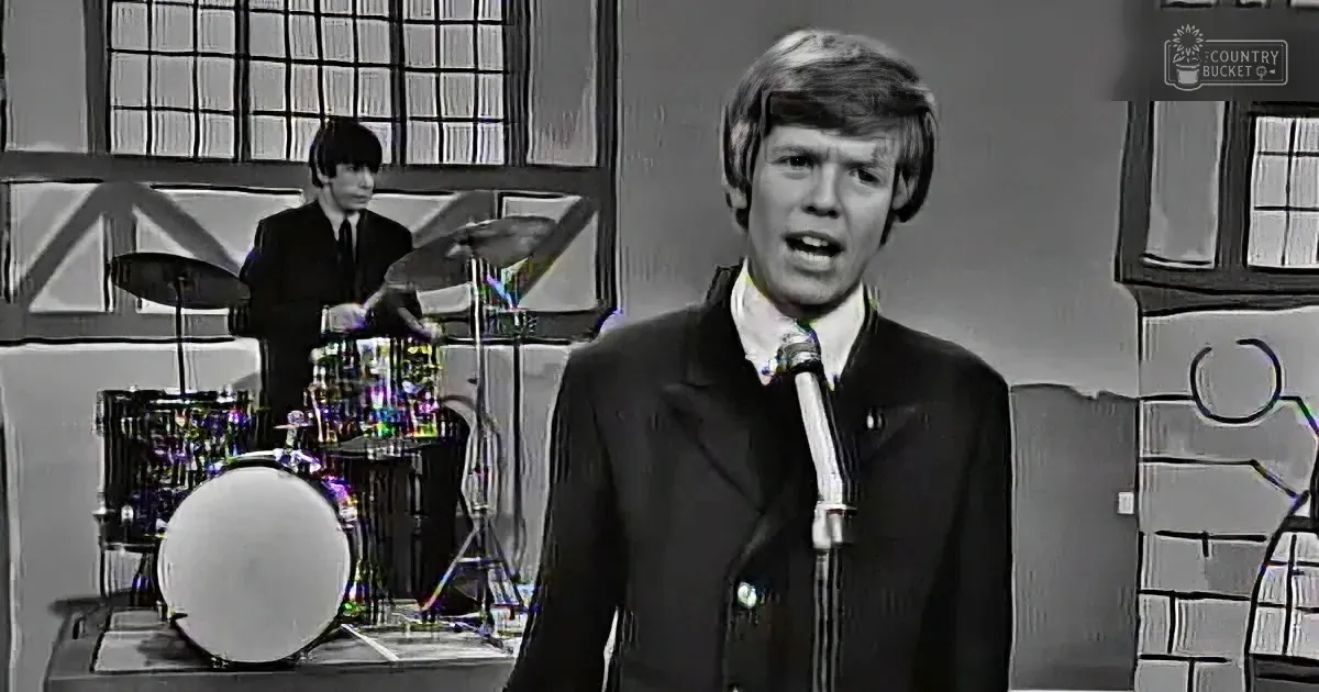 A man in a suit sings into a microphone, with a drummer in the background and a stylized backdrop.