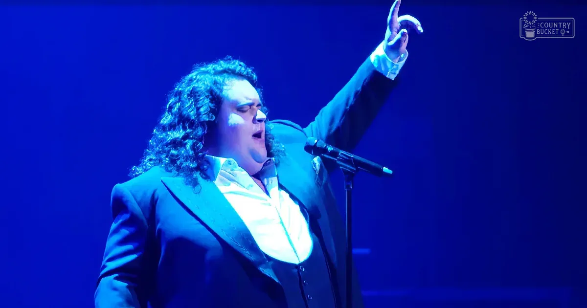 An adult male performer in a dark suit jacket over a white shirt with rolled-up sleeves, wearing a ring on his raised left hand, performing on stage under blue lighting.