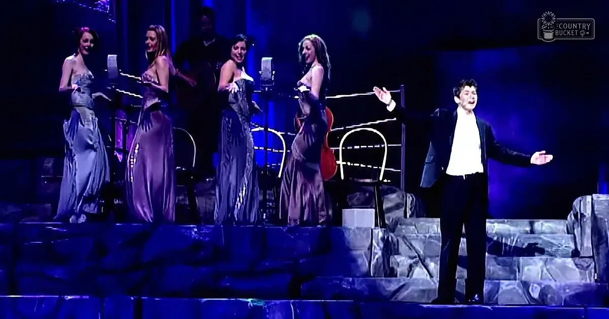 A man in a suit sings passionately on stage with four women in elegant dresses, accompanied by a guitarist.