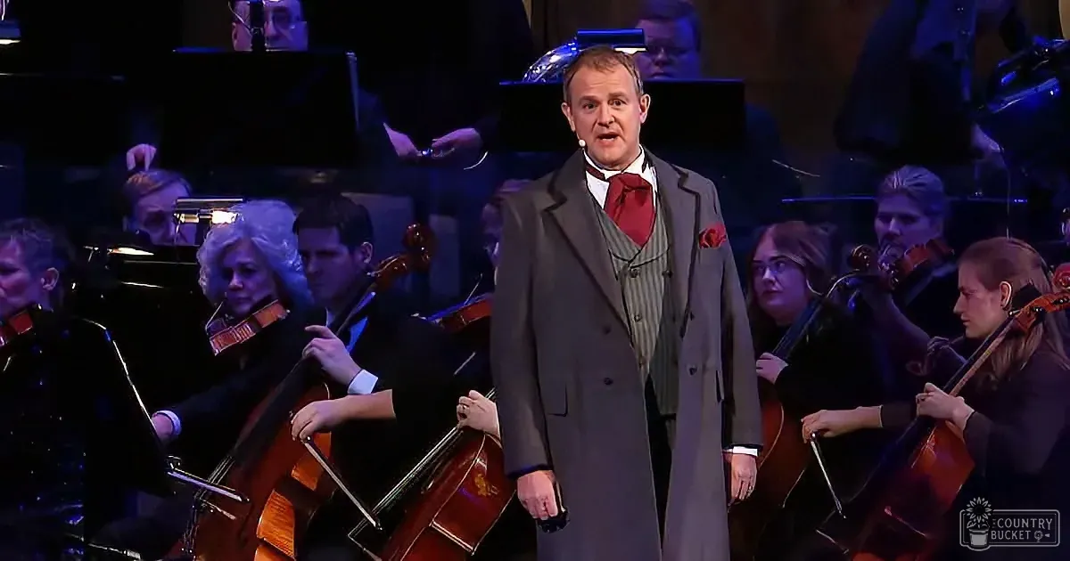 A man in a suit sings on stage with an orchestra playing in the background.