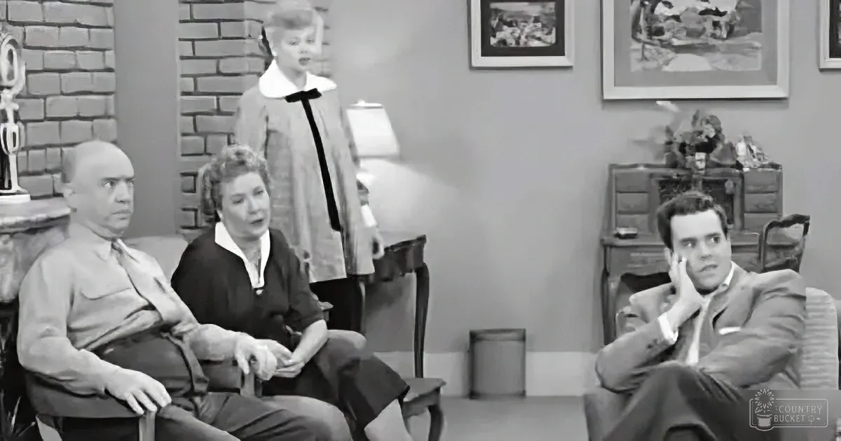 A man in a suit sits in a chair, hand on face, while others watch in a living room setting.