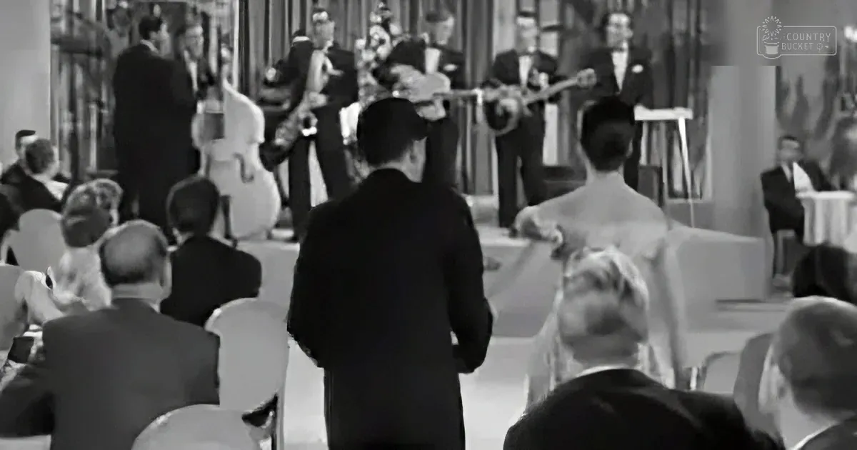 A man in a suit stands in front of a seated audience, facing a band playing in a formal setting.