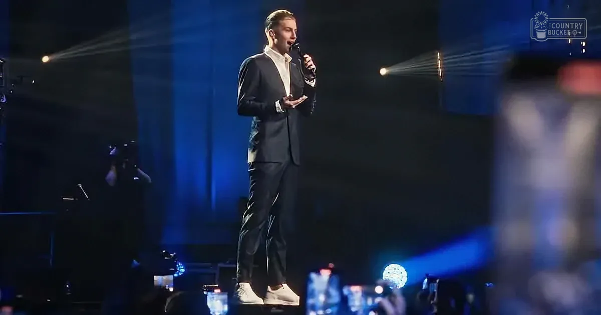 A man in a suit stands on stage holding a microphone, illuminated by stage lights.