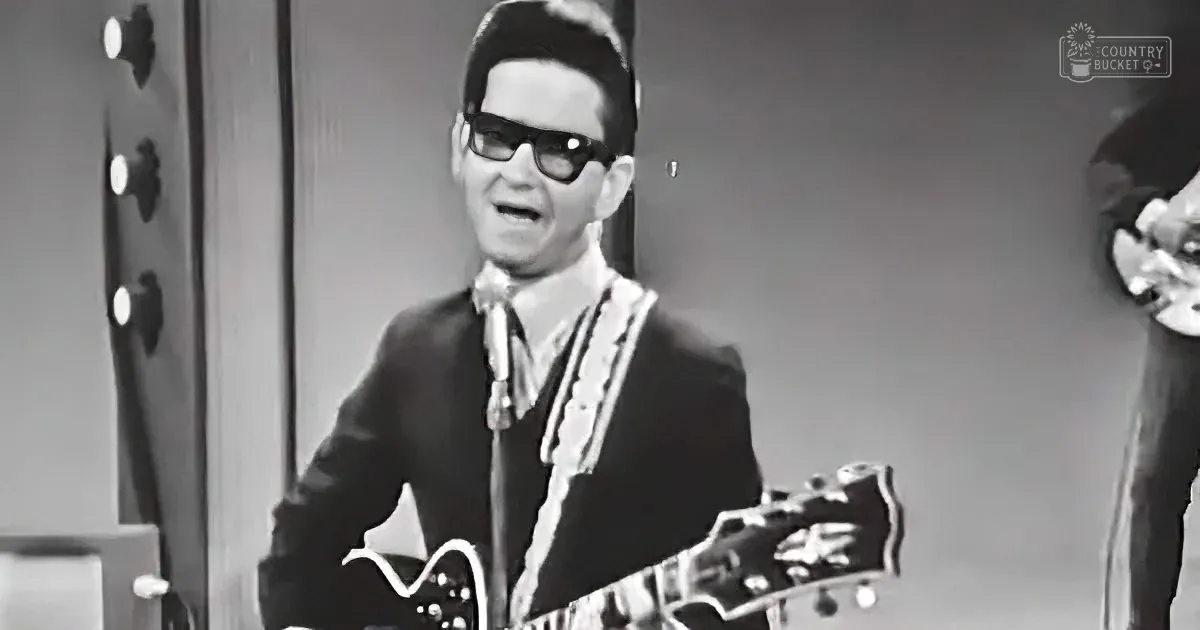 A man in a suit and sunglasses plays a guitar, wearing a white scarf, in a black-and-white image.