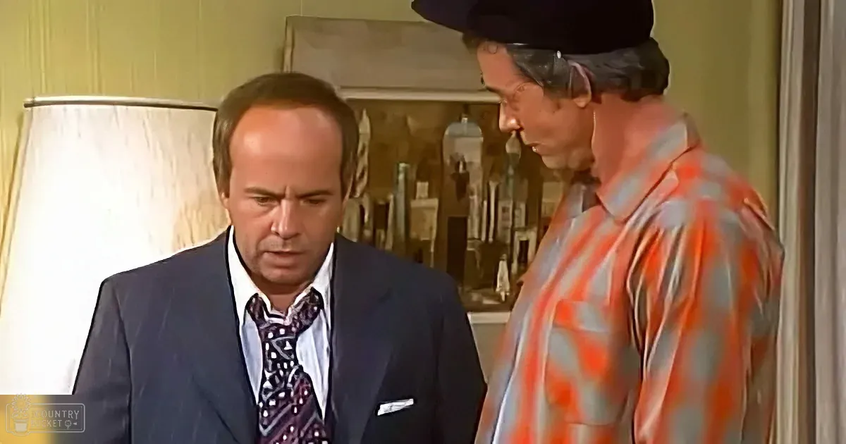 A man in a suit and tie looks down while another man in a striped shirt and hat stands beside him.
