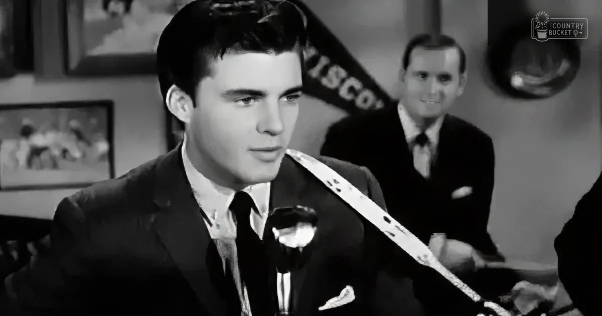 A man in a suit and tie speaks into a microphone, with another man in the background.