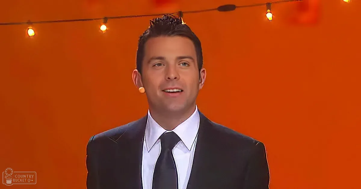 A man in a suit and tie stands against an orange backdrop with string lights.