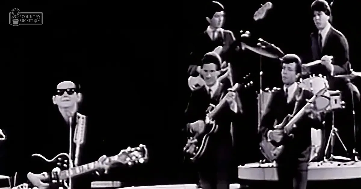 A man in sunglasses plays guitar on stage with a band, including a drummer and a guitarist.