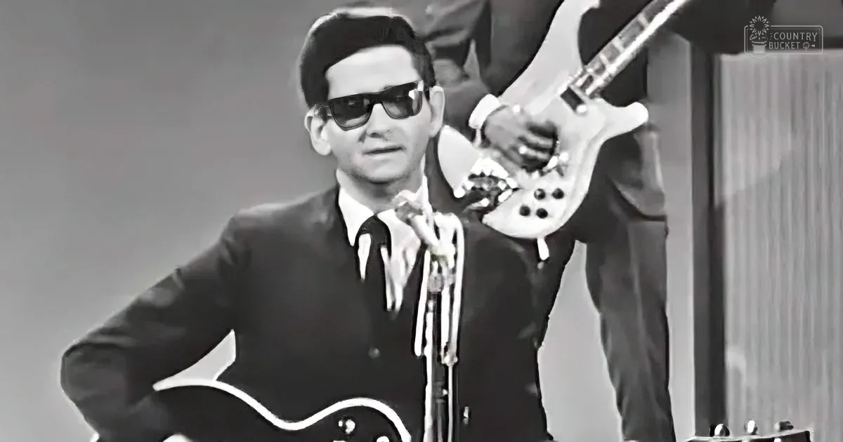 A man in sunglasses and a suit plays a guitar on stage, with another musician in the background.