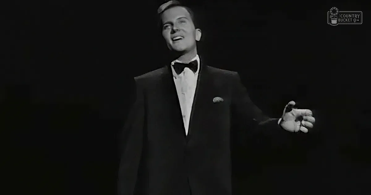 A man in a tuxedo and bow tie is singing, holding a microphone, against a dark background.