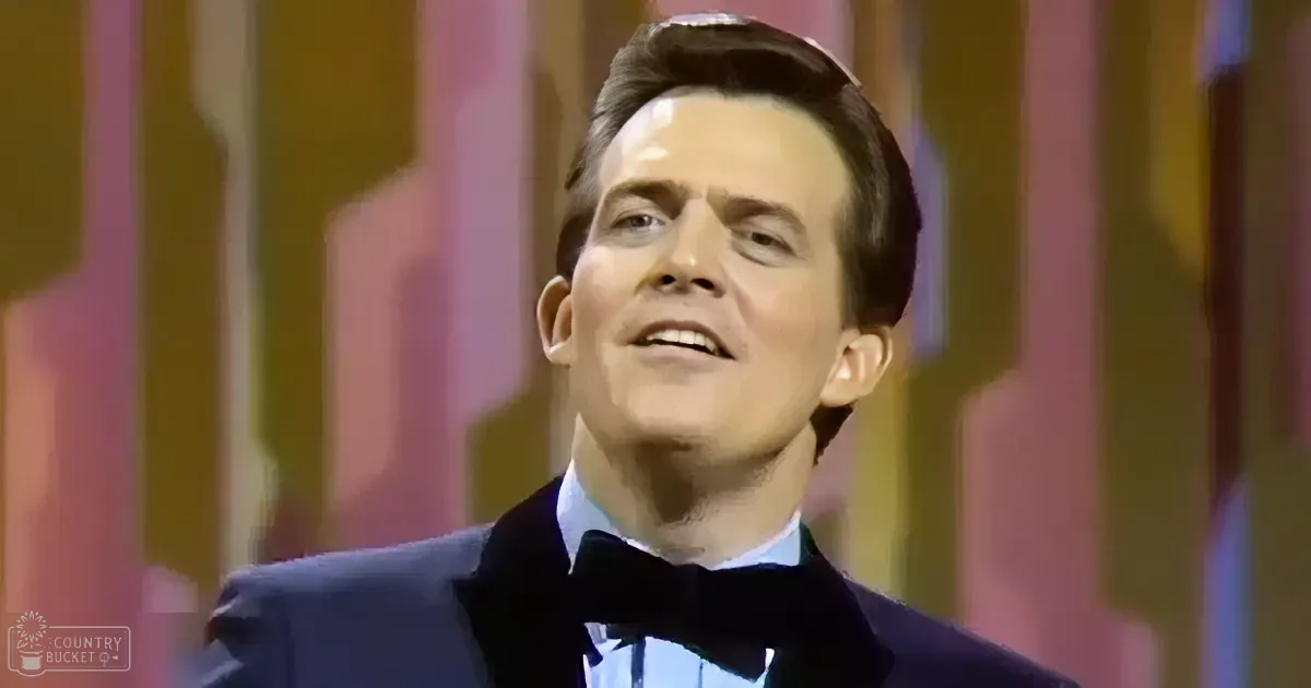 A man in a tuxedo with a bow tie, smiling, against a colorful striped background.