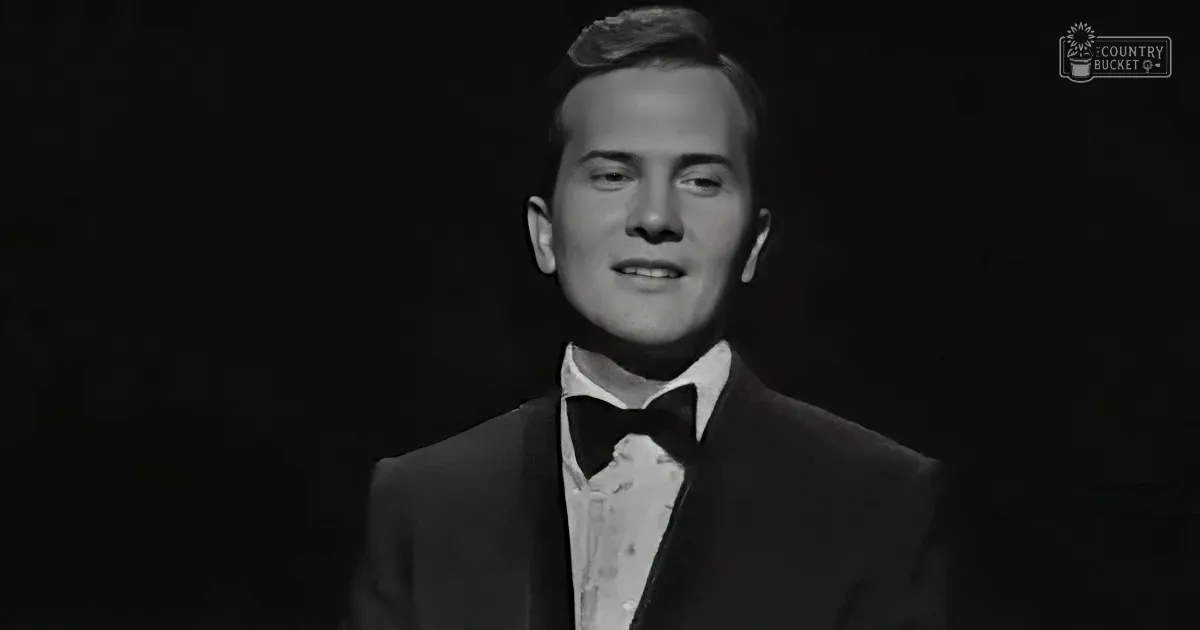 A man in a tuxedo and bow tie stands against a dark background, smiling.