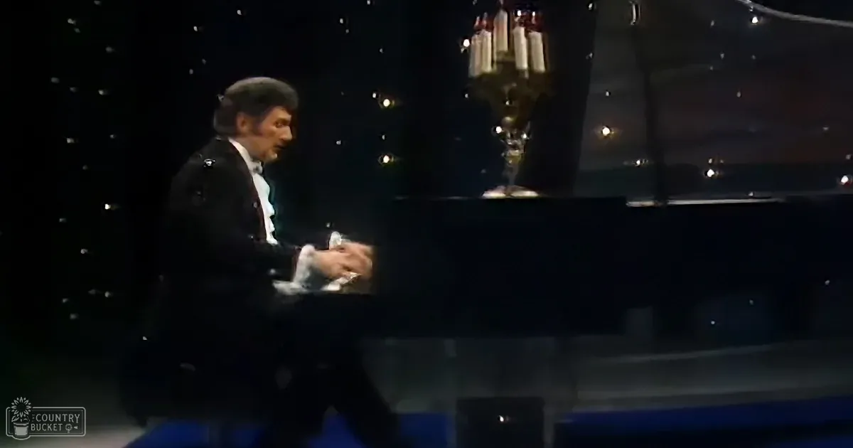 A man in a tuxedo plays a grand piano on a stage with a chandelier.