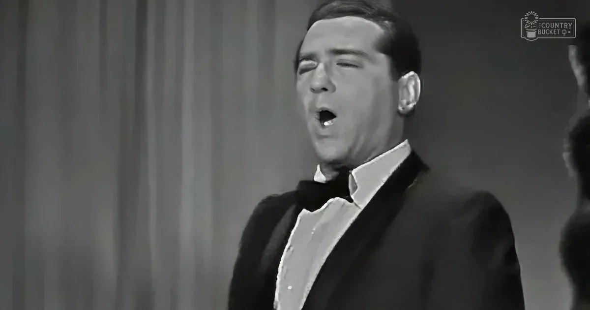 A man in a tuxedo sings passionately, mouth open, against a curtain backdrop.