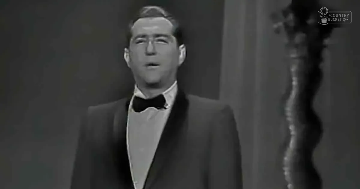 A man in a tuxedo sings on stage, mouth open, with a dark background and a trophy-like object to the side.