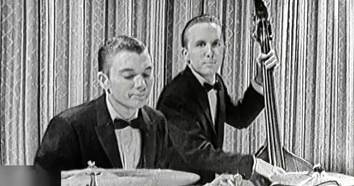 A man in a tuxedo sits at a table, smiling, while another man plays a double bass in the background.