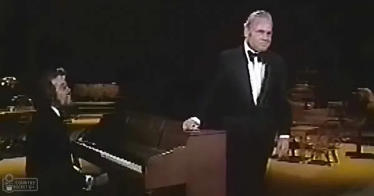 A man in a tuxedo stands beside a seated pianist in a dimly lit stage setting.