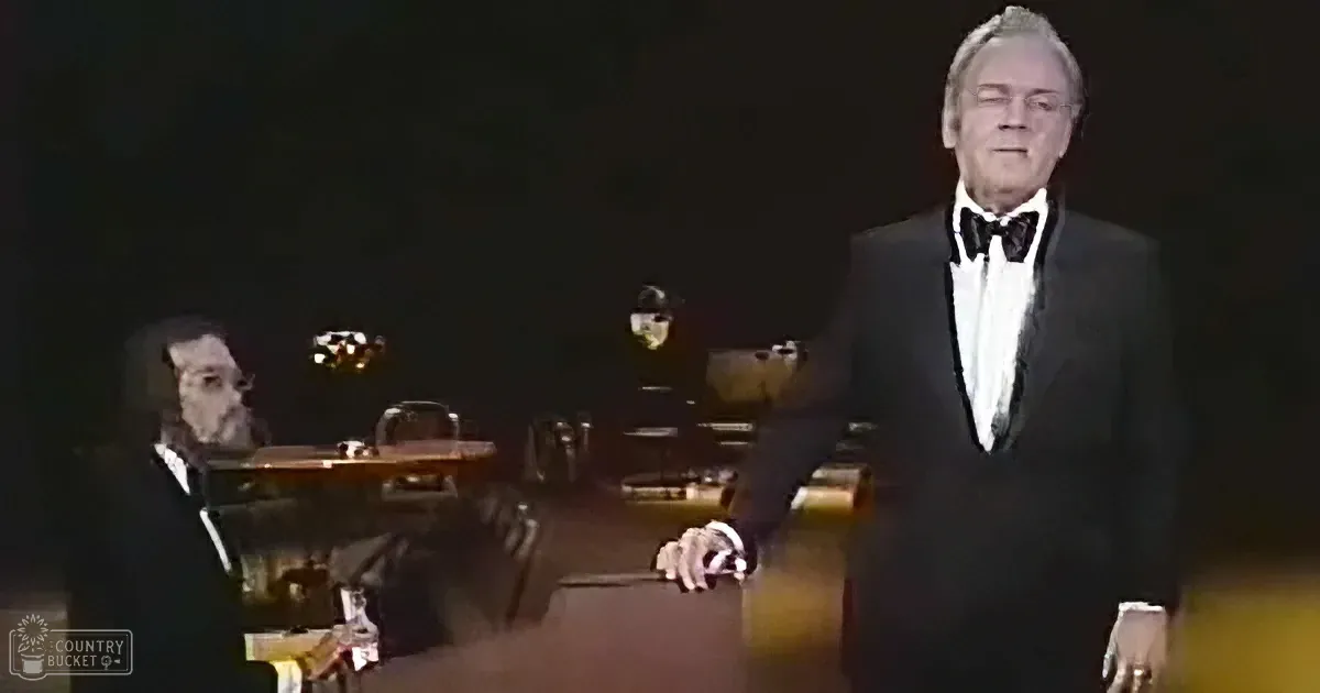 A man in a tuxedo stands in a dimly lit room, with another man seated at a piano in the background.