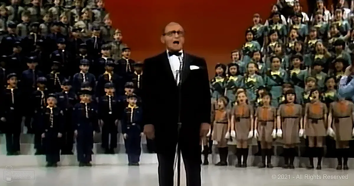 A man in a tuxedo stands in front of a choir, singing, with a large audience in the background.